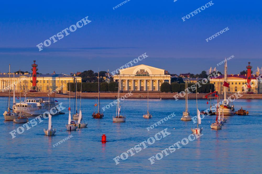 На фотографии изображены яхты и катера, дрейфующие на водах Невы перед Стрелкой Васильевского острова. В центре композиции выделяется здание Биржи, обрамленное Ростральными колоннами. Тёплый свет летнего вечера подчеркивает элегантность архитектуры и красоту городской панорамы. Этот вид является одним из символов Санкт-Петербурга.